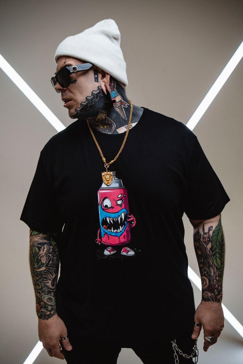 A male model stands confidently in front of a beige background, with illuminated light poles forming an "X" shape behind him. He wears a black oversized T-shirt featuring a colorful graphic of a cartoon character, paired with black pants adorned with chains. His look is completed with a white beanie, sunglasses, and noticeable tattoos on his arms. The overall vibe reflects a bold urban streetwear style.