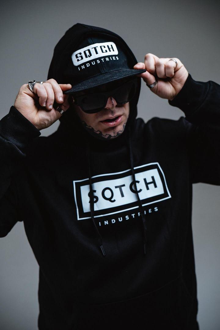 A male model adjusts the brim of his black cap while wearing a matching black hoodie that features the logo "SQTCH INDUSTRIES." He sports sunglasses and has visible tattoos on his arms. The background is a muted beige, enhancing his edgy urban style. His expression is confident and focused.