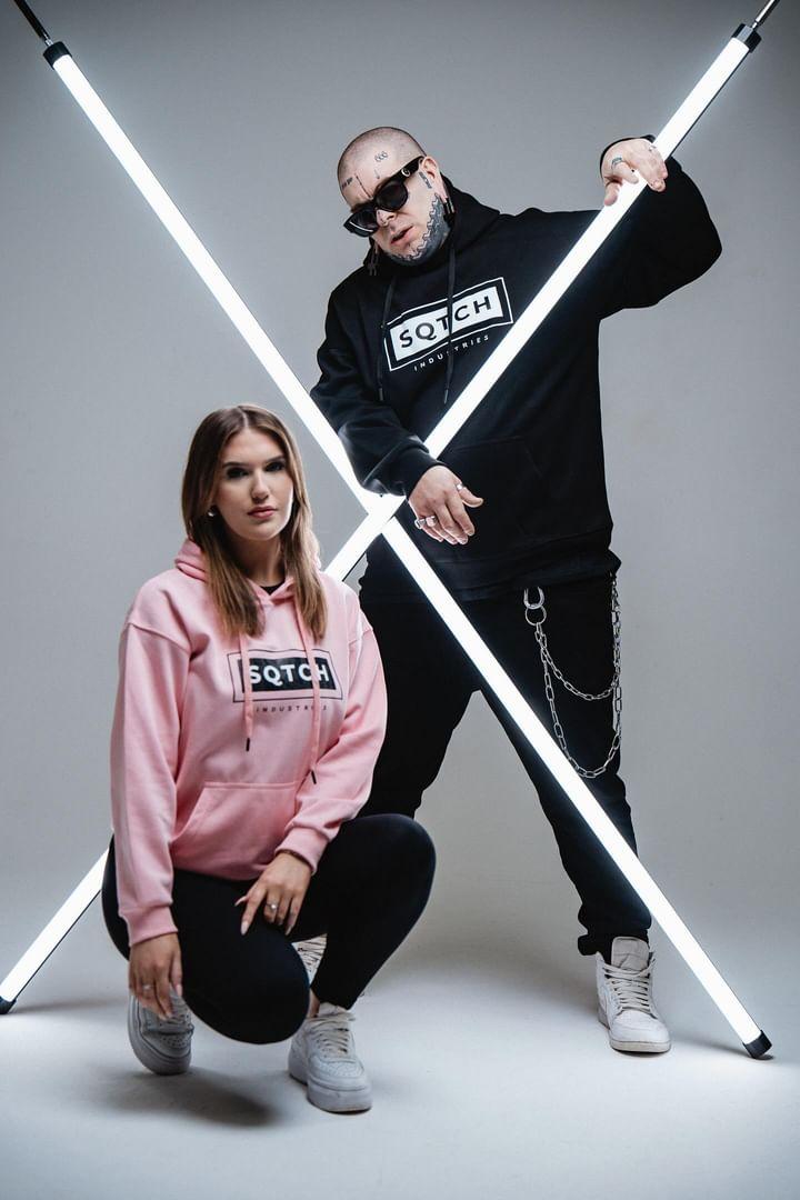 A male and female model pose against a gray background, creating a striking composition with illuminated light poles arranged in an "X" shape. The female model, wearing a pink hoodie and black leggings, kneels with her hood up, while the male model stands confidently beside her in a black hoodie, accessorized with sunglasses and chain details. Both models are styled in urban fashion, showcasing a modern streetwear aesthetic.