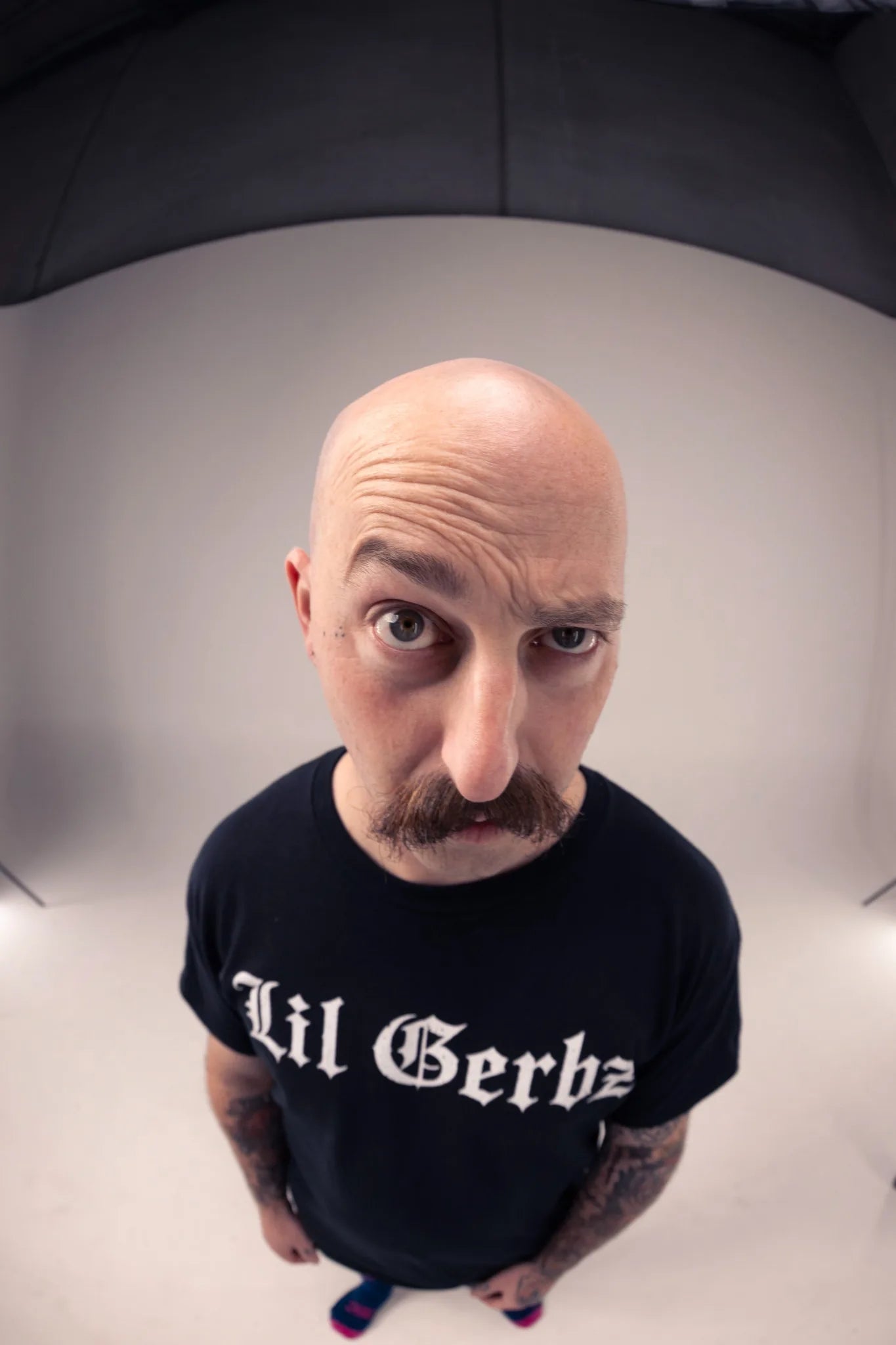 A man with a mustache looking up at the camera, wearing a black t-shirt with 'Lil Gerbz' printed in white gothic lettering, photographed in a studio setting with a fisheye lens.