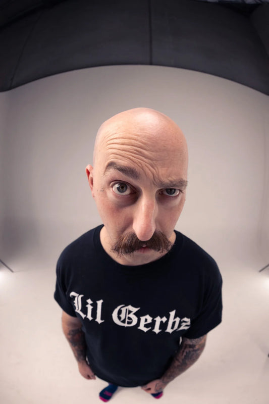 A man with a mustache looking up at the camera, wearing a black t-shirt with 'Lil Gerbz' printed in white gothic lettering, photographed in a studio setting with a fisheye lens.