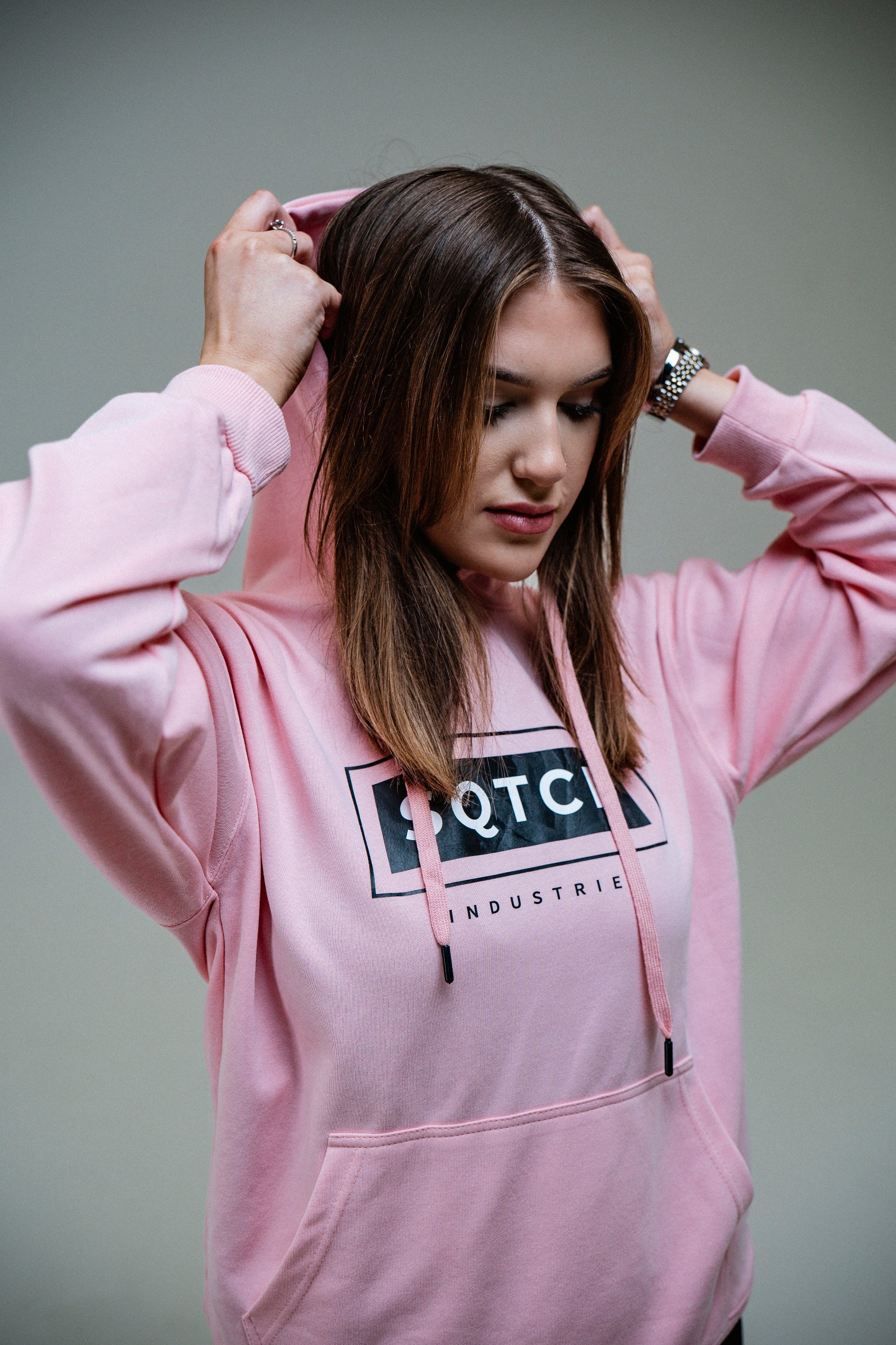 A female model pulls the hood of her pink sweatshirt over her head, creating a focused expression. She stands against a soft beige background, and the sweatshirt features a logo with the word "SQTCH" prominently displayed. Her hair falls straight and she wears a watch on her wrist, contributing to a stylish urban look.