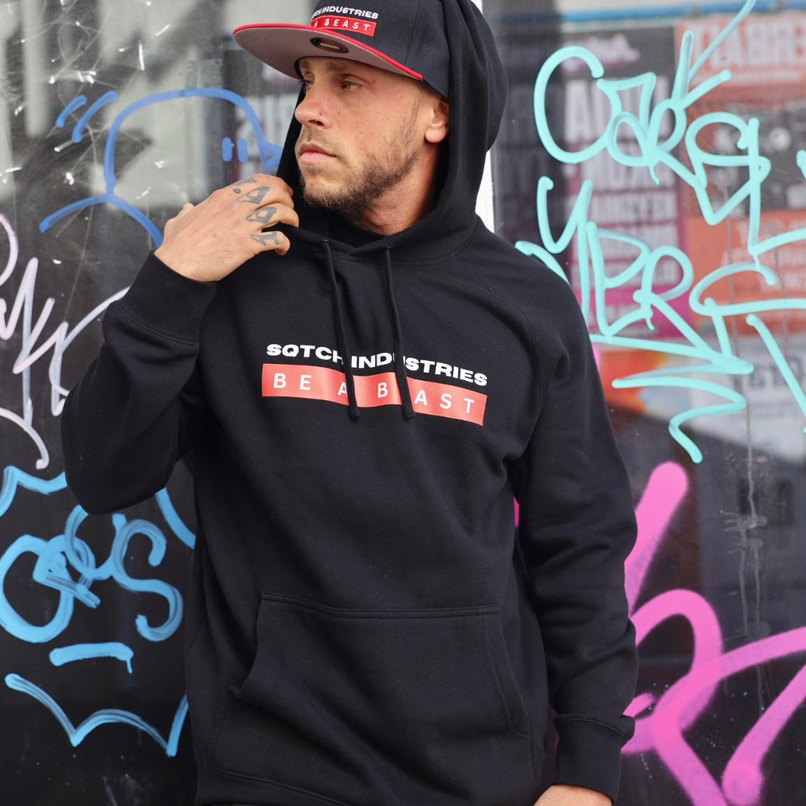 Model wearing Sqtch Industries black “Be A Beast” hoodie in urban graffiti street setting